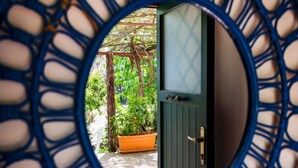 Une vue à travers une fenêtre circulaire à cadre bleu, qui révèle une porte verte et un mur blanc avec une fenêtre.