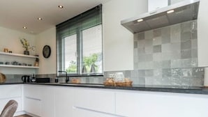 A modern kitchen with white cabinets, a black countertop, and a window with blinds. - Holiday Home in Texel Near the Sea (De Koog)