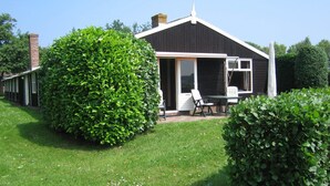 Ein eingeschossiges Haus mit dunkler Fassade, ein großer, grüner Busch davor und eine Terrasse mit Tisch und Stühlen.