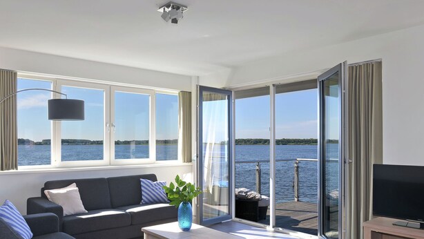 A modern living room with a sofa, a coffee table, a TV, and large windows offering a view of the water. - Apartment in Kamperland by Veerse Meer Lake (Kamperland)