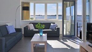 A modern living room with a sofa, a coffee table, and a view of a body of water through large windows. - Apartment in Kamperland by Veerse Meer Lake (Kamperland)