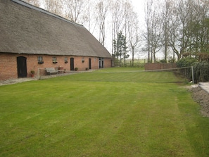 A tennis court with a net, a brick building with a thatched roof, and a grassy area. - Rural Holiday Home With Outdoor Pool & Sauna (Zuidzande)