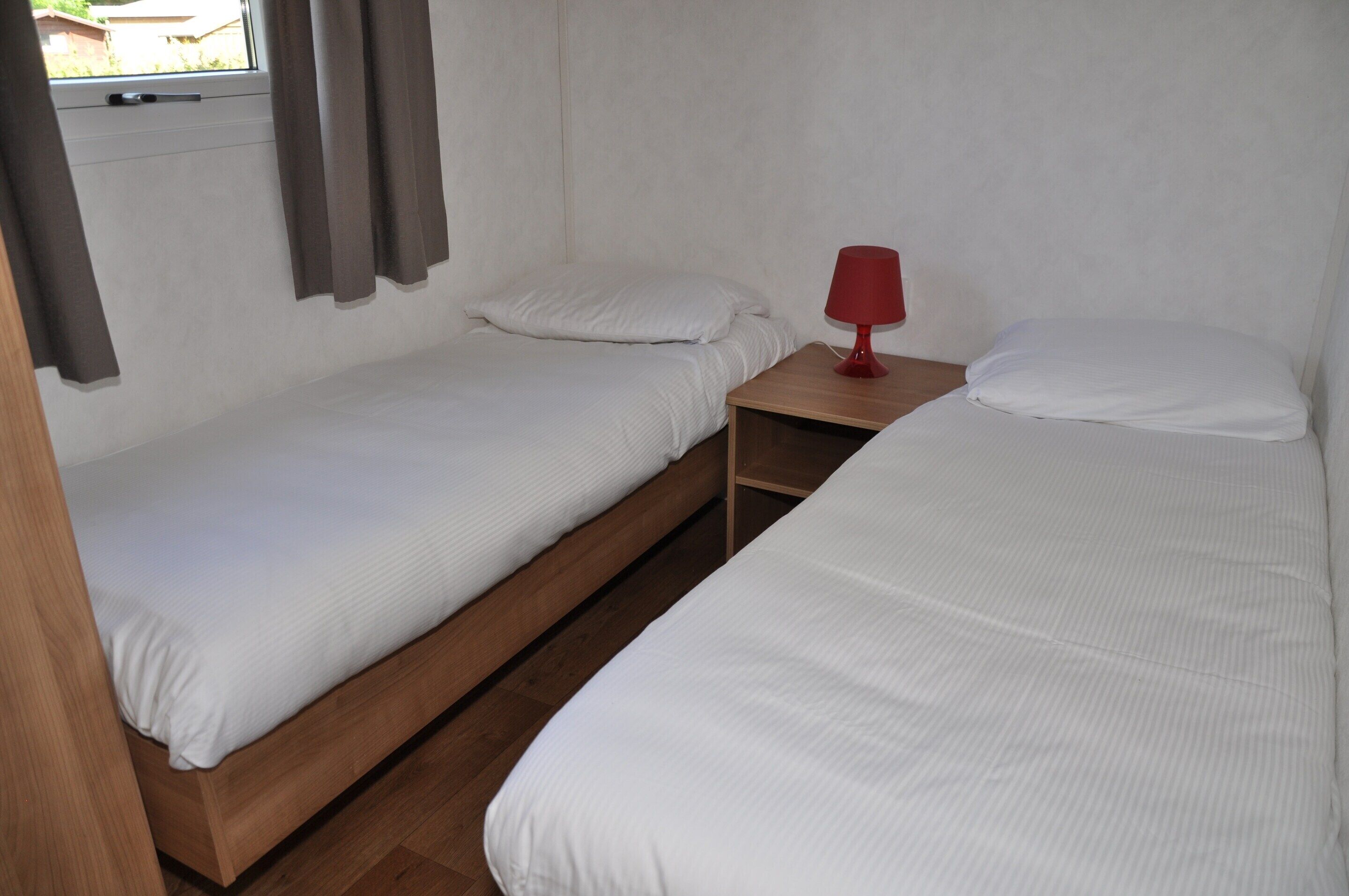 A room with two single beds, a wooden nightstand with a red lamp, and a window with curtains.