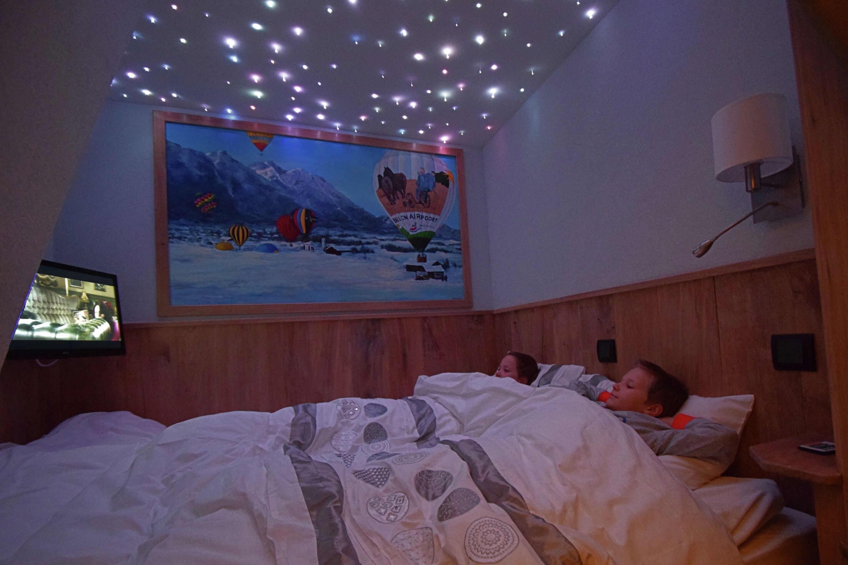 A bedroom with a bed, a television on the wall, a painting of a hot air balloon, and a ceiling with lights.