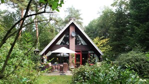 A triangular wooden building with a red door, surrounded by lush greenery and a patio area with outdoor furniture. - Spacious Holiday Home in Eersel With Open Fire (Eersel)