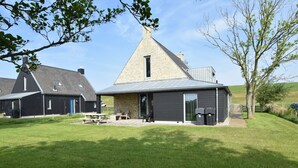 Een modern gebouw met een puntig dak, een terras met buitenmeubels en een groot raam.
