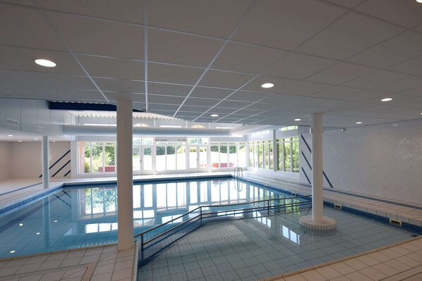 Une piscine intérieure avec des parois en verre transparent, un sol carrelé de carreaux blancs et des luminaires au plafond.
