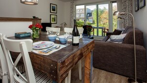 A dining area with a wooden table, chairs, and a bottle of wine. There is a window with a view of houses and trees. - Apartment in Hollum Near the Sea (Hollum)