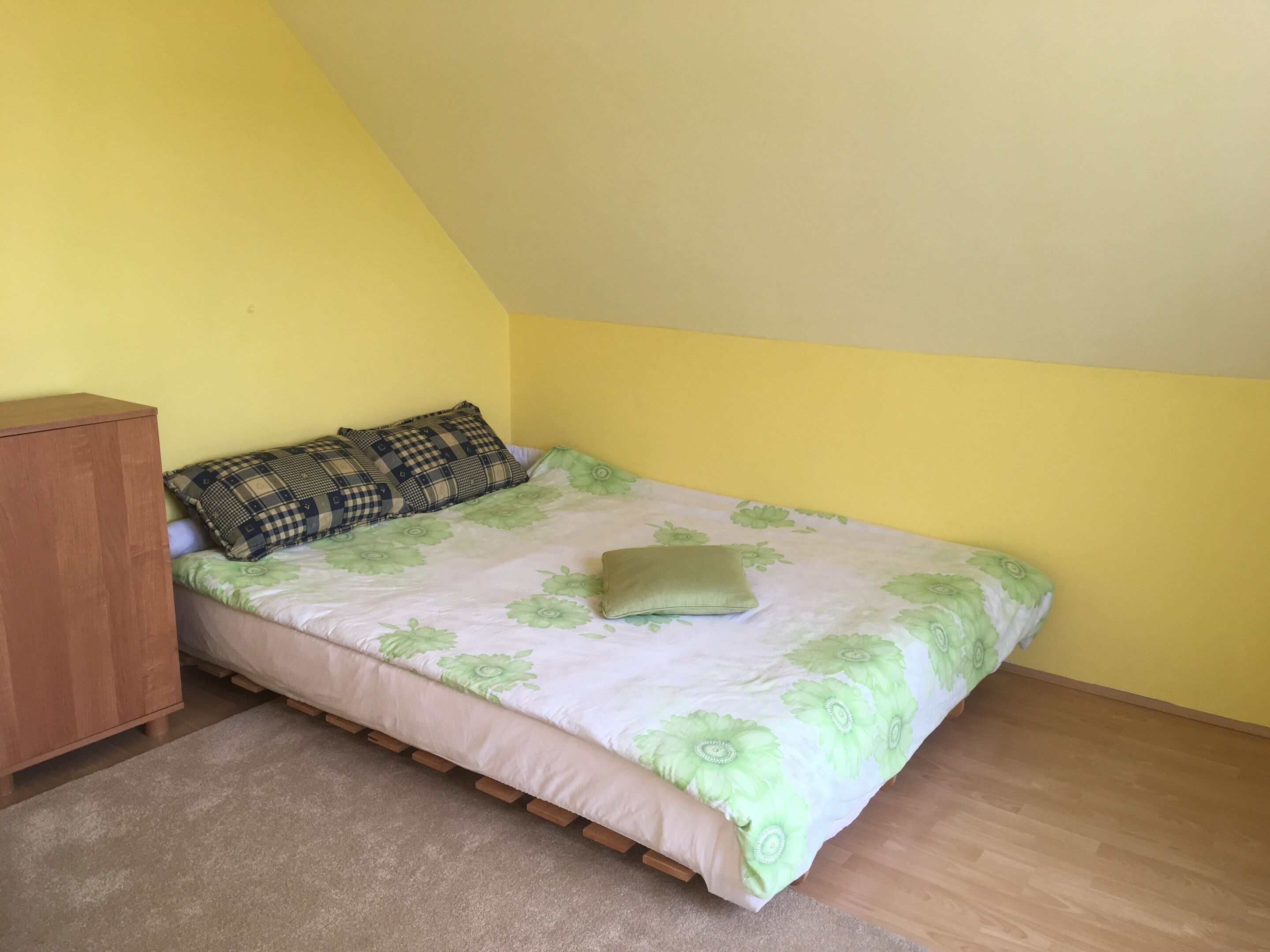 Una habitación pequeña con una cama individual, suelo de madera, pared amarilla y un armario de madera.