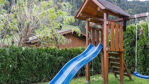Een houten speelconstructie met een blauwe glijbaan en schommel, in een gazon met een houten hek en bomen op de achtergrond.