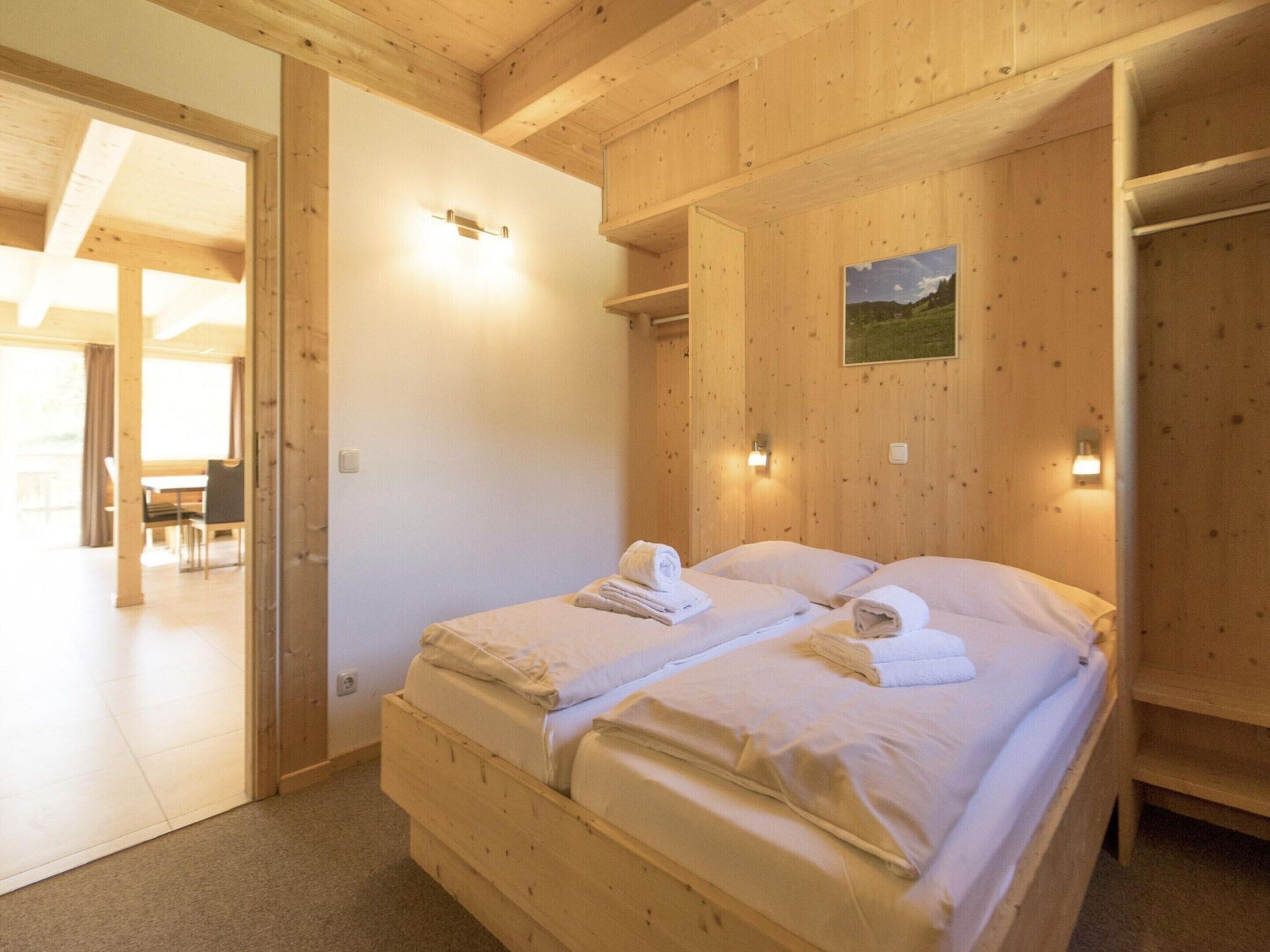 A wooden bedroom with two beds, a small painting on the wall, and a dining area visible through an open door.