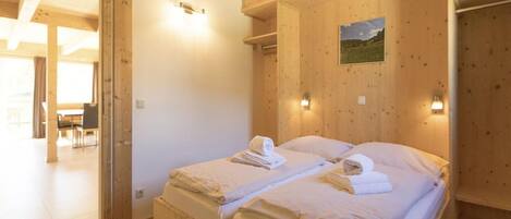 Een houten slaapkamer met twee bedden, een klein schilderij aan de muur, en een eethoek zichtbaar door een open deur.