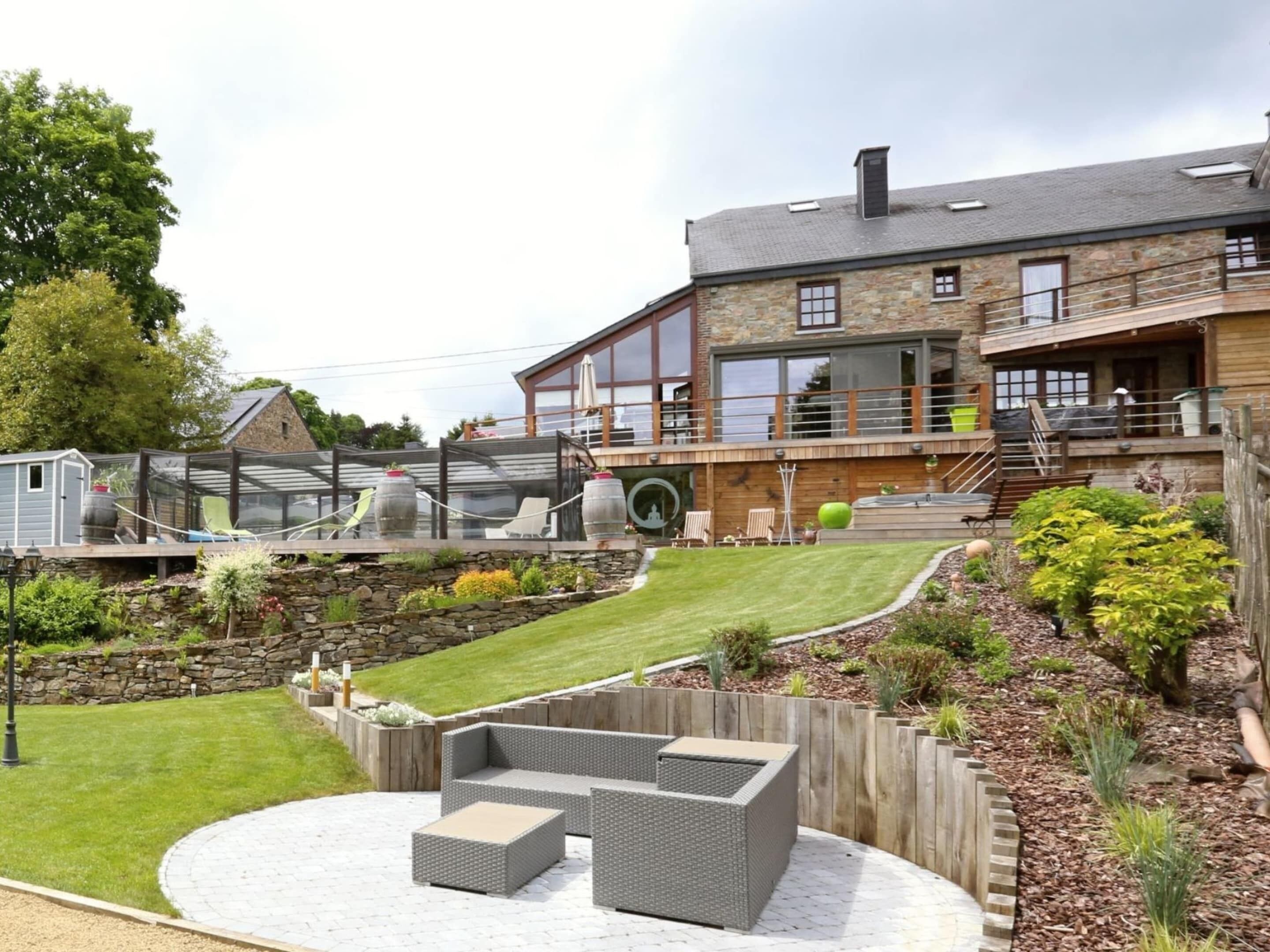 Un bâtiment moderne avec une façade en pierre, un espace extérieur aménagé avec des places assises couvertes et un jardin bien entretenu.