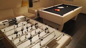 Una mesa de billar, una mesa de futbolín al frente, un sofá y una ventana con persianas.