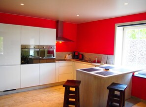 A modern kitchen with white cabinets, a red accent wall, a built-in oven, and a window with a view of greenery. - Spacious Home With Games (Ellezelles)