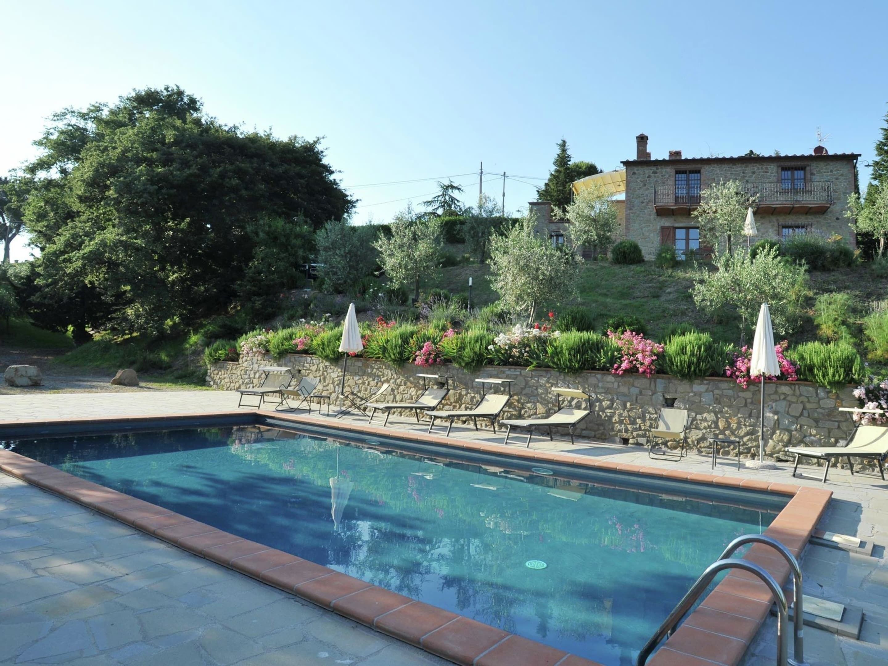 Une piscine entourée d’un mur de pierre et d’une végétation luxuriante, avec des chaises longues et des parasols à proximité.