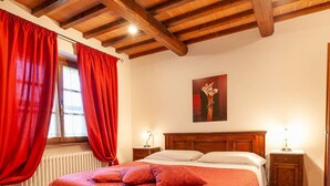A bedroom with a wooden headboard, a bed with red bedding, two bedside tables with lamps, and a window with red curtains. - Villa in Lisciano Niccone With Pool & Nature (Lisciano Niccone)