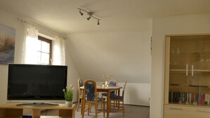 Olohuone, jossa on televisio, puinen ruokapöytä, tuoleja ja lasiovilla varustettu kaappi.