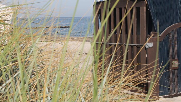 A beach hut with a striped awning, situated on a sandy beach with grass in the foreground. - Ferienhaus in Bastorf for 5 Personen (Bastorf)