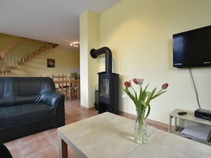 A living room with a black sofa, a table with a vase of flowers, a black wood-burning stove, a TV mounted on the wall, and a staircase in the background.