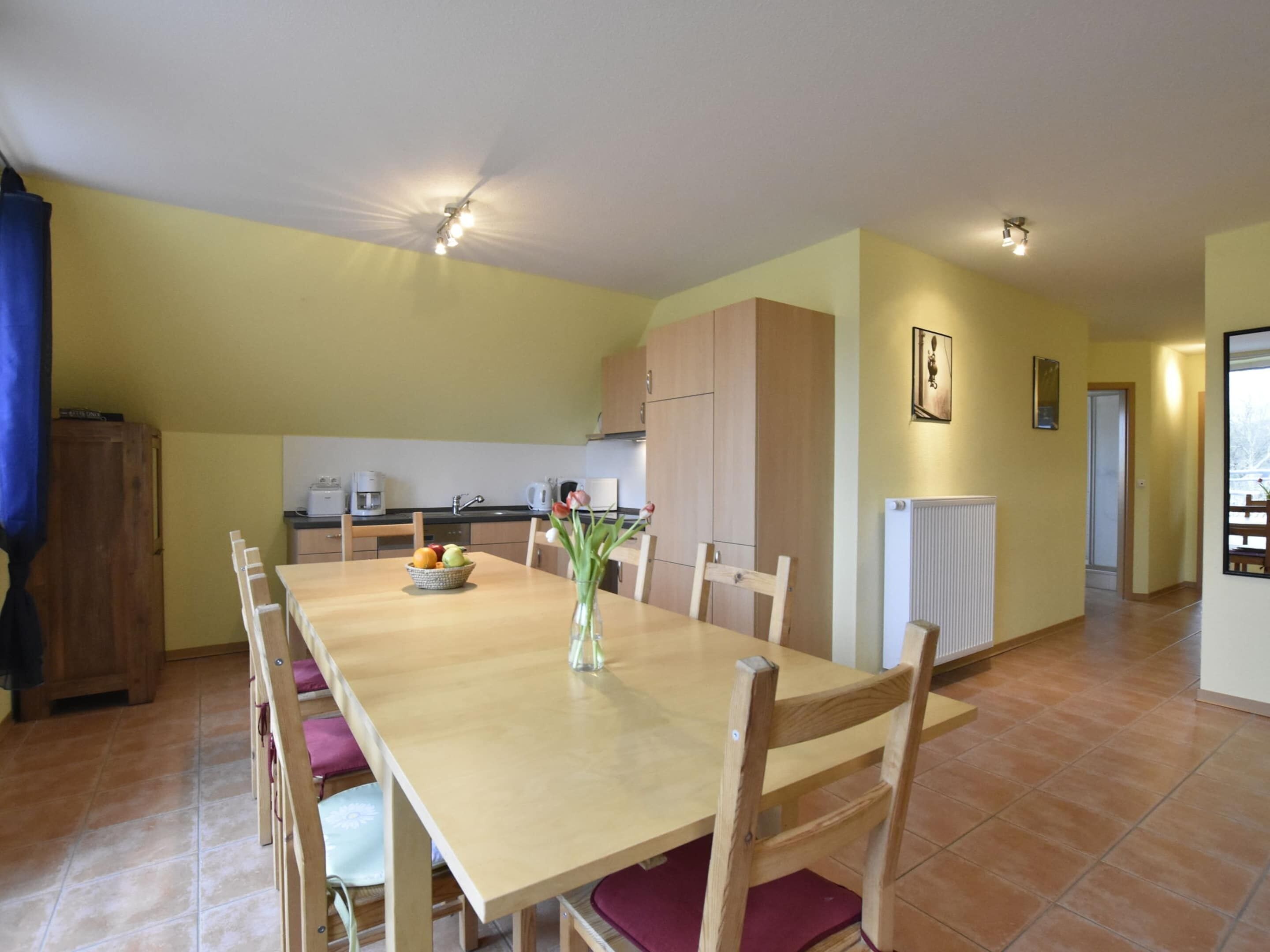 A dining area with a long table, chairs, a vase with flowers, and a kitchen in the background.