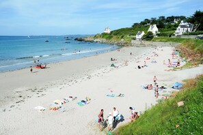 En strand med folk som solar och badar, några parasoller och hus på en kulle i bakgrunden.