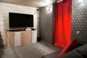 A room with a flat-screen TV, a wooden cabinet, a red curtain, patterned wallpaper, and a black sofa.