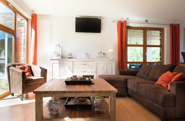 A living room with a wooden coffee table, a sofa, a chair, a TV mounted on the wall, and a window with curtains.