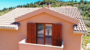 Una casa con techo de tejas, un balcón con contraventanas de madera y vistas a una ladera boscosa.