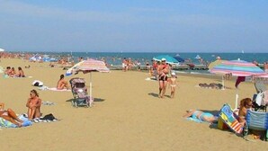 Ein voller Strand mit Menschen, die sonnenbaden und spazieren gehen, bunten Sonnenschirmen und einem klaren Himmel.