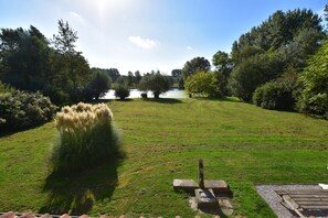 A grassy area with a wooden bench, a stone structure, and a body of water in the background. - Holiday Home in Saint-omer Near Lake & Forest (Saint-Omer)