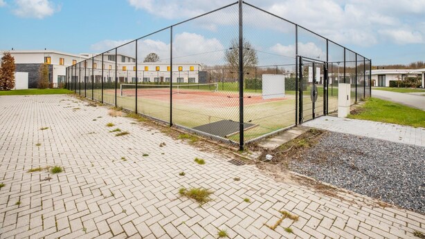 A sports facility with a fenced tennis court, a brick pathway, and modern buildings in the background. - Modern Villa with Large Garden by the Water with Hot Tub & Infrared Sauna (Zeewolde)