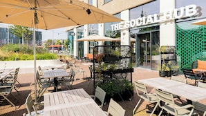 Un espace extérieur aménagé avec des tables et des chaises, un grand parasol, et un bâtiment sur lequel est apposée une enseigne indiquant : « THE SOCIAL HUB ».