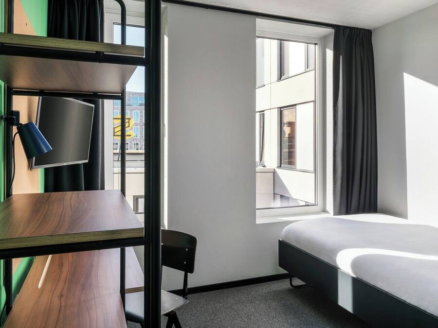 Et kompakt hotelværelse med køjeseng, skrivebord med stol, et fladskærms-tv og et stort vindue med udsigt over byen.
