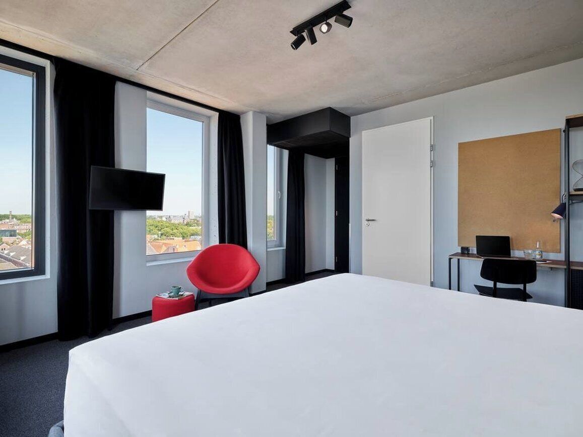 A hotel room with a large bed, a red chair, a desk with a computer, a flat-screen TV mounted on the wall, and a view of the city through large windows.
