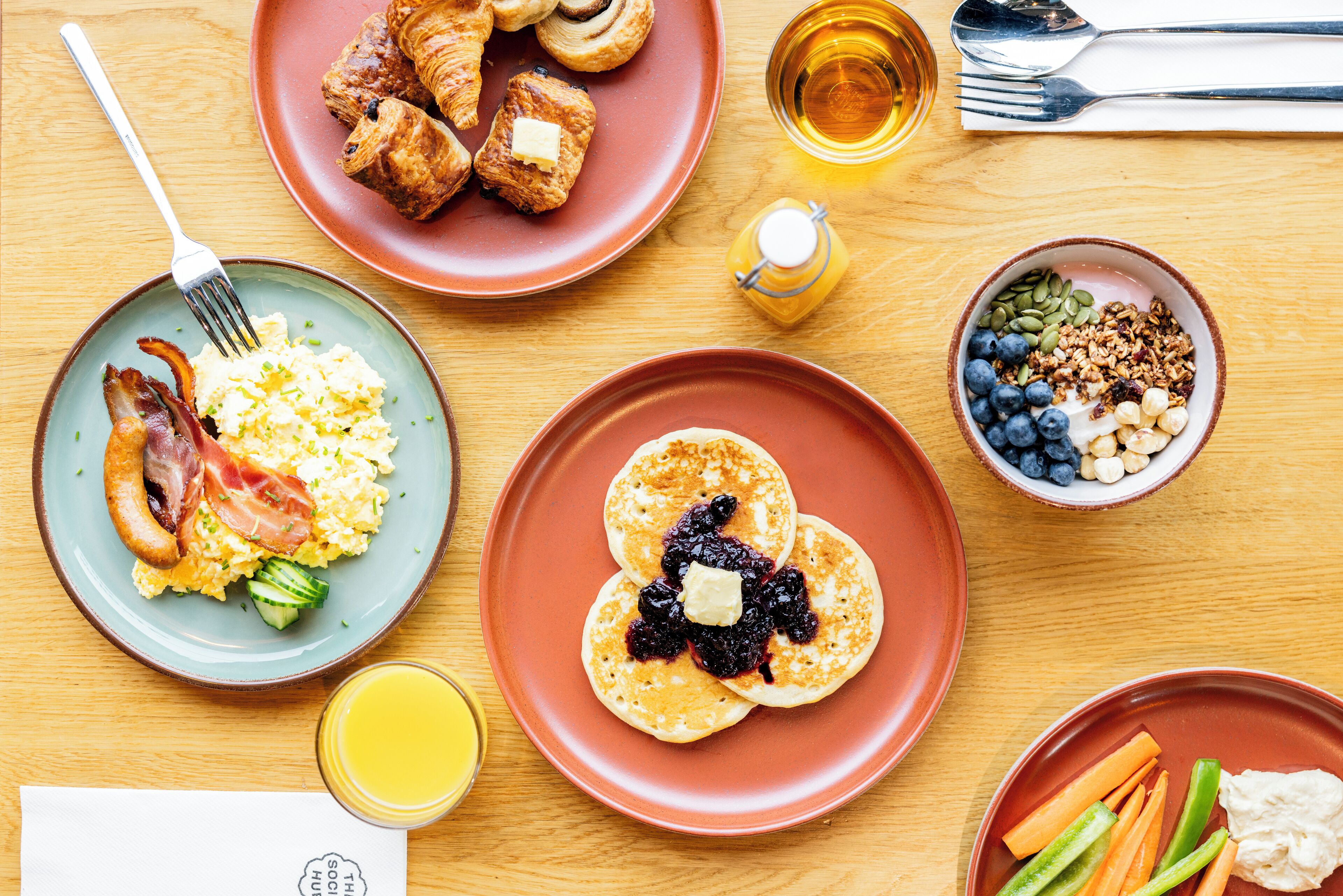 Impostazione colazione con diversi piatti, tra cui uova, pancetta, pancake e una ciotola di frutta e granola.