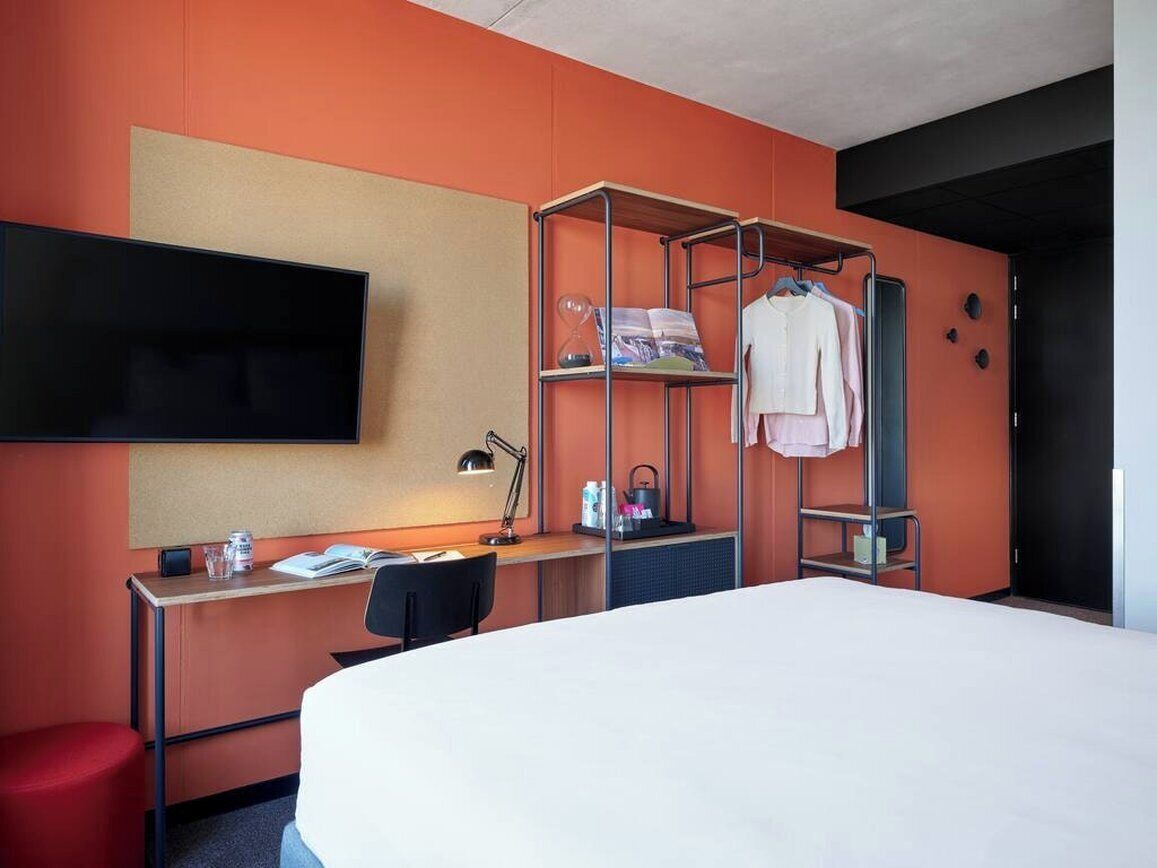 A hotel room with a bed, a desk, a TV mounted on the wall, a shelf with clothes, and a red stool.