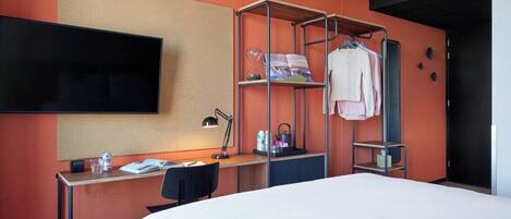 A hotel room with a bed, a desk, a TV mounted on the wall, a shelf with clothes, and a red stool.