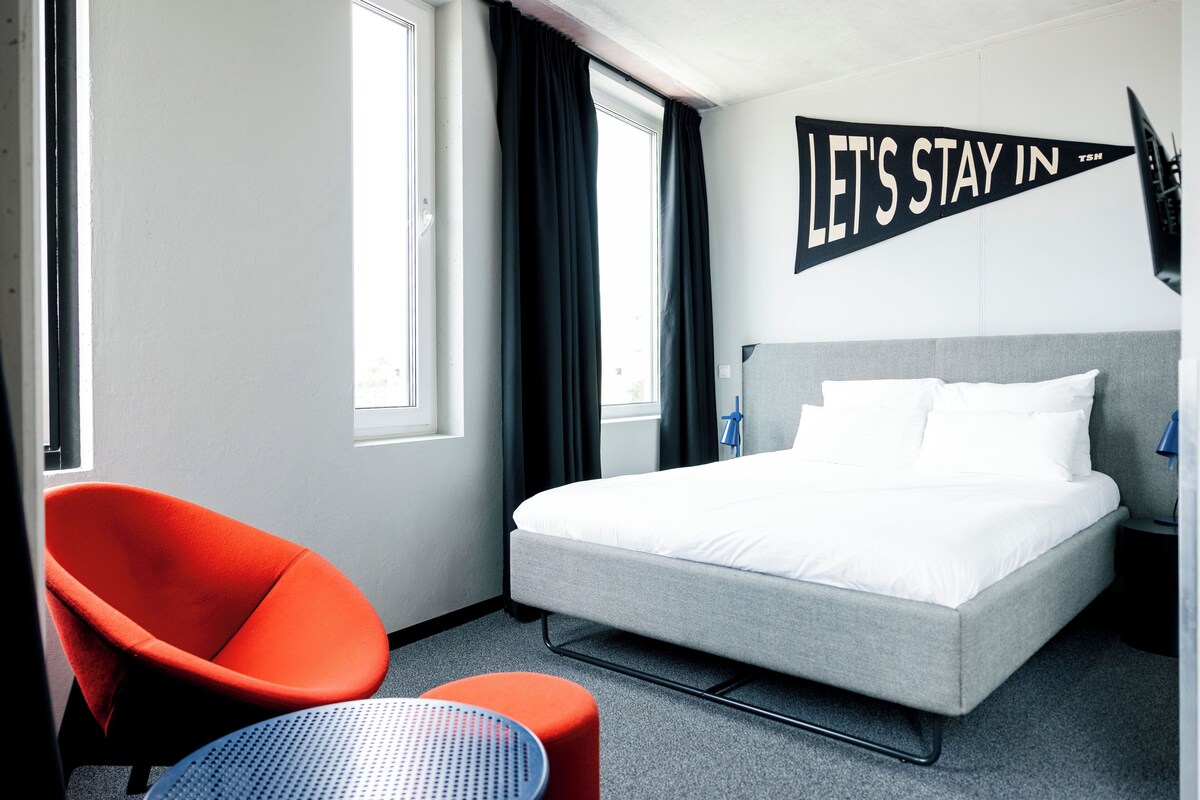 Una habitación de hotel con una cama, una silla roja, una mesita y un letrero que dice 'LET'S STAY IN'.
