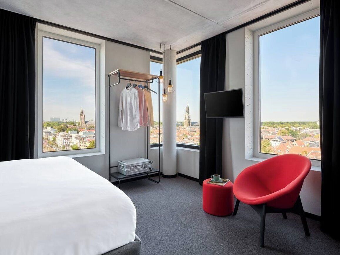 A hotel room with a bed, a red chair, a TV, and a view of a cityscape.