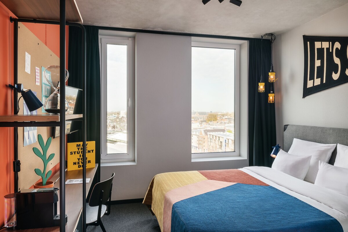 Habitación de hotel con cama, dos ventanas, escritorio con lámpara, silla y un tablón de anuncios con un cartel.