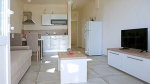A modern kitchen with white cabinets, a microwave, and a refrigerator. There is a wooden table with a bowl and a plant. A beige sofa and a TV are also visible. - Apartment in Ljubac Near the Sea (Razanac)