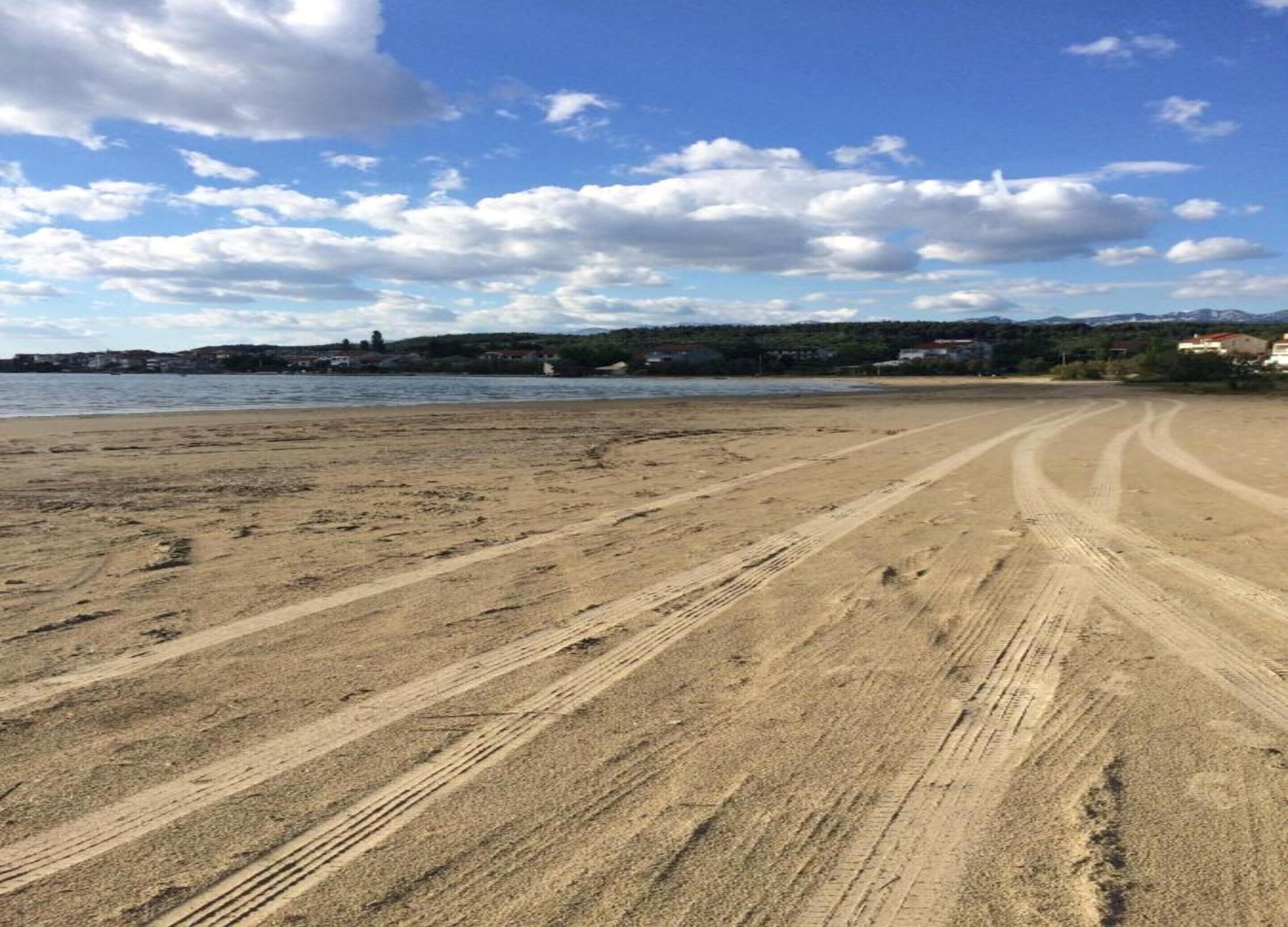 A wide sandy beach with tire tracks, a clear sky, and distant buildings.