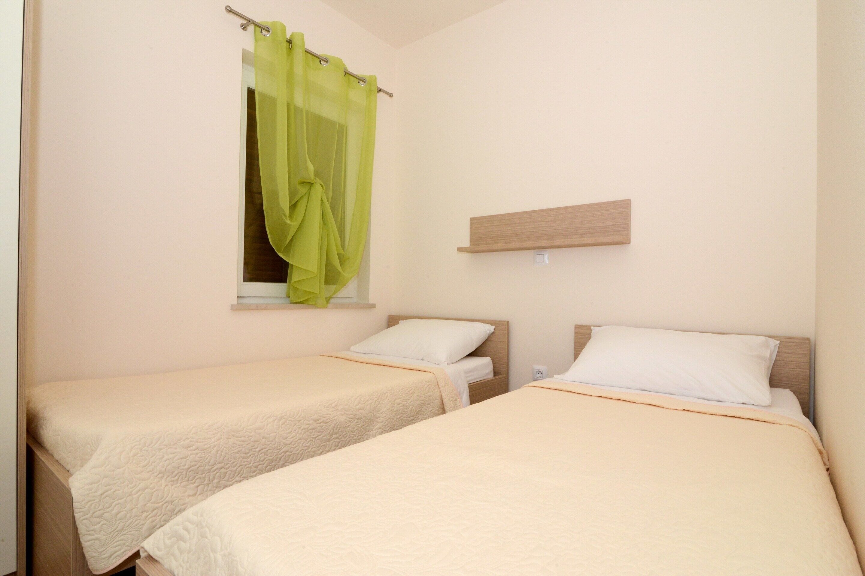 Una habitación con dos camas individuales, una ventana con cortina translúcida, un estante de madera y una pared de color claro.