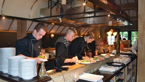 A kitchen with chefs preparing food, a counter with plates and a wine glass, and shelves with bottles. - Cozy Holiday Home with Dishwasher near Zwolle (Dalfsen)