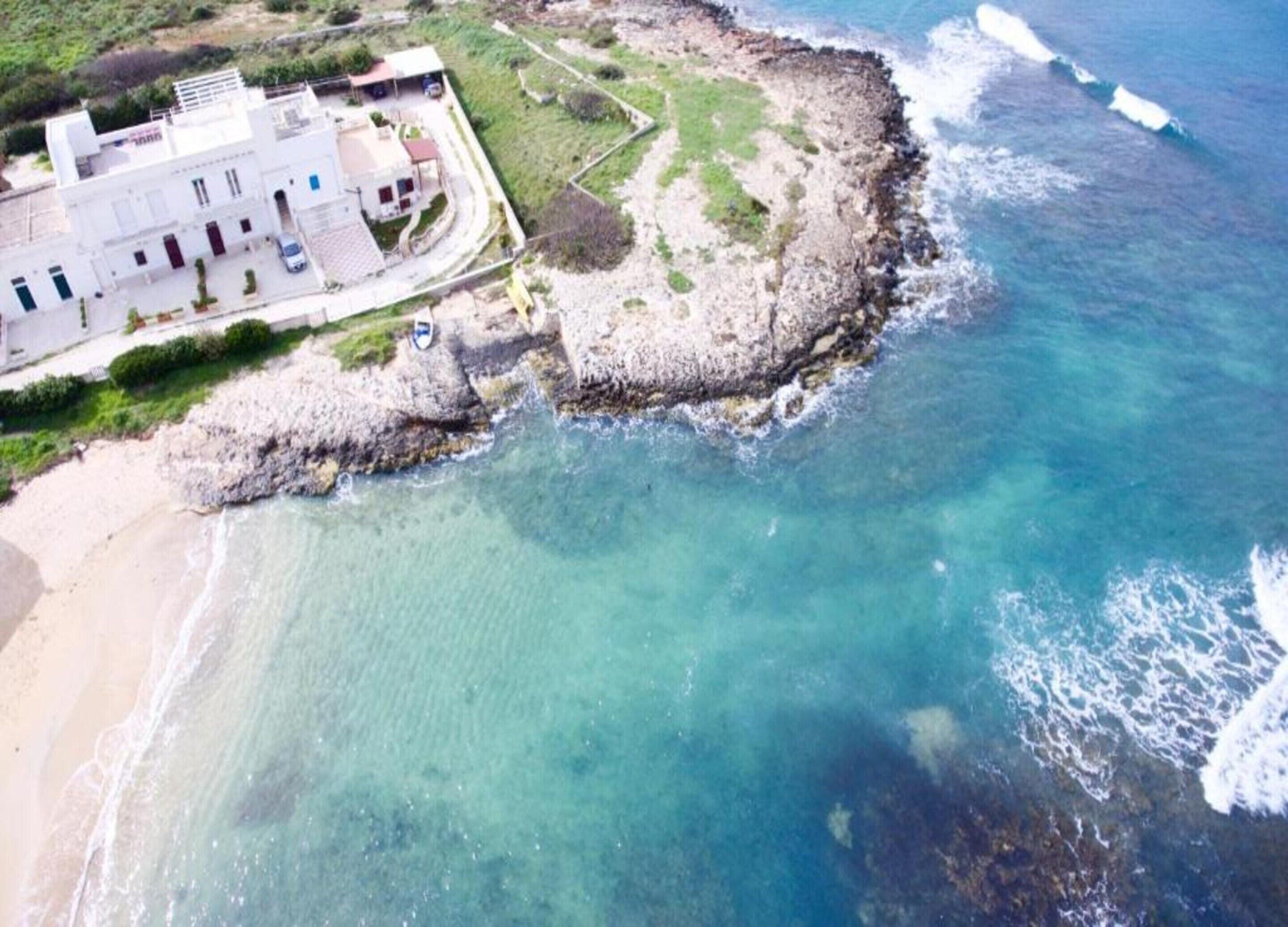 An aerial view of a coastal property with a building, a rocky shoreline, and clear blue waters.
