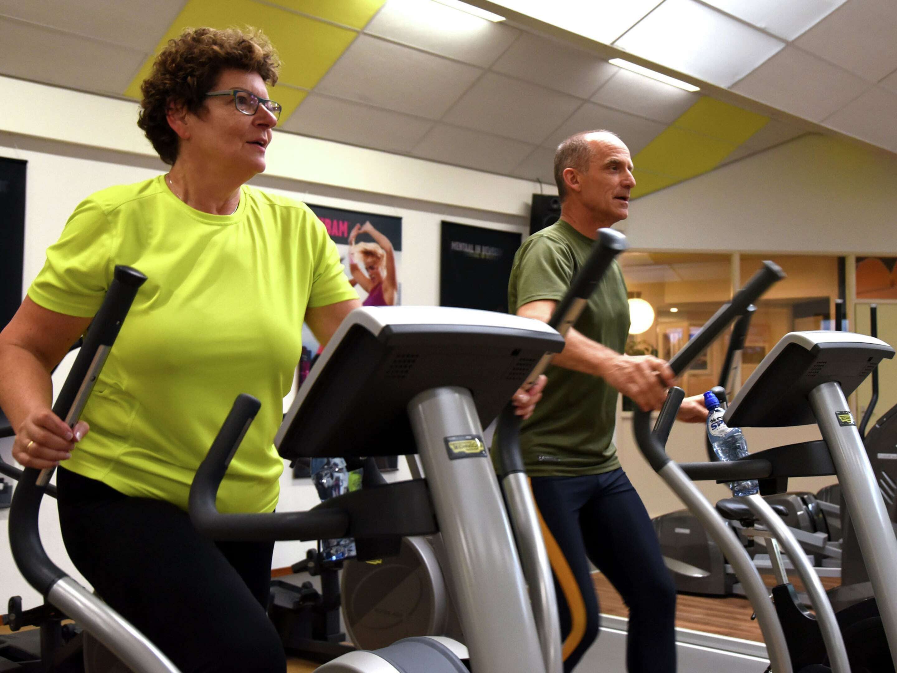 Two people using treadmills in a gym with exercise equipment and a poster on the wall.