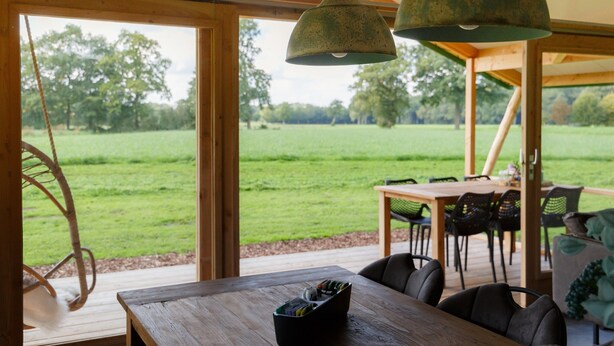 A wooden dining table with chairs, a hanging light fixture, and a view of a grassy field through large windows. - Tent Lodge With Dishwasher in Twente (Haaksbergen)