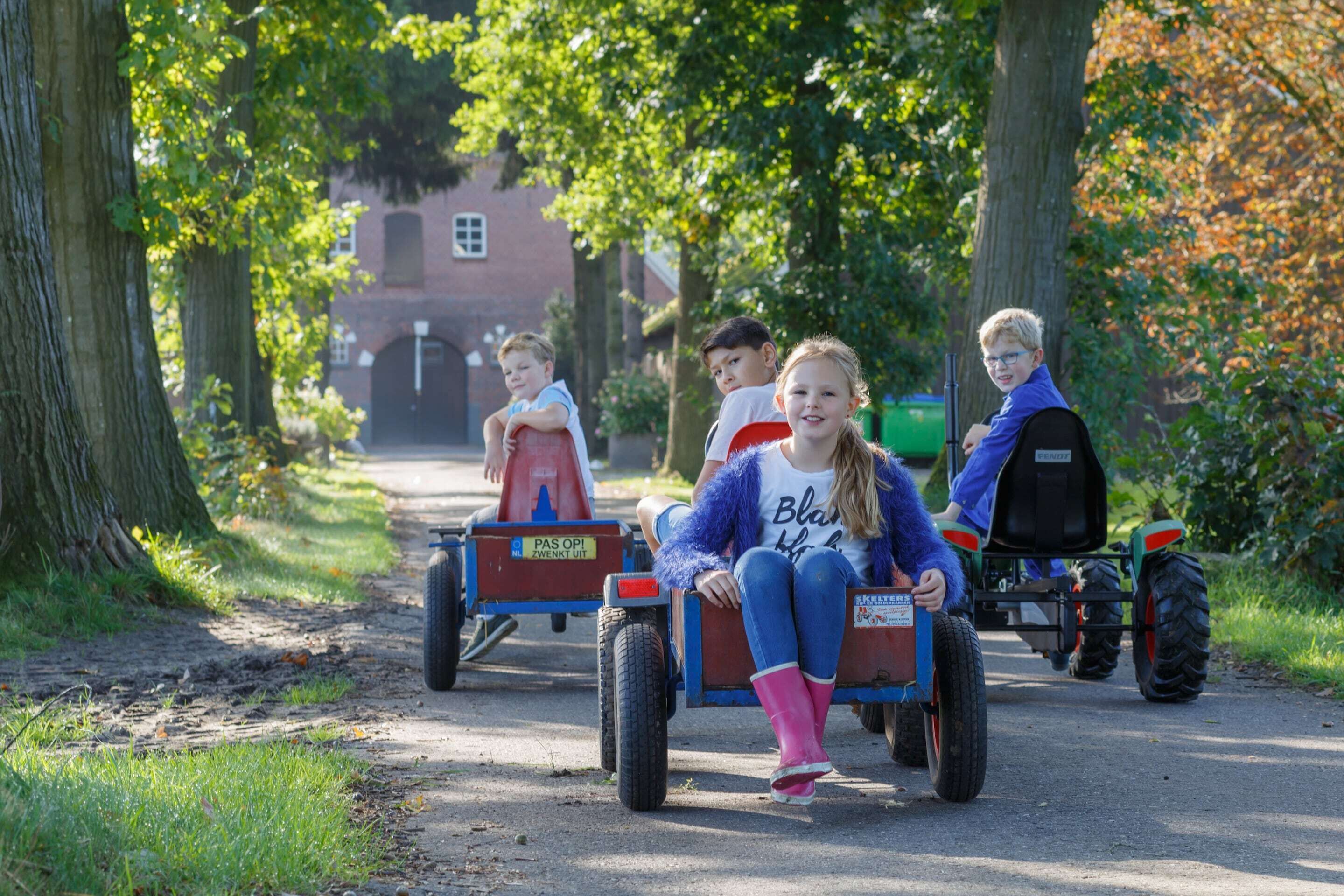 Children riding a small vehicle on a paved path surrounded by trees.