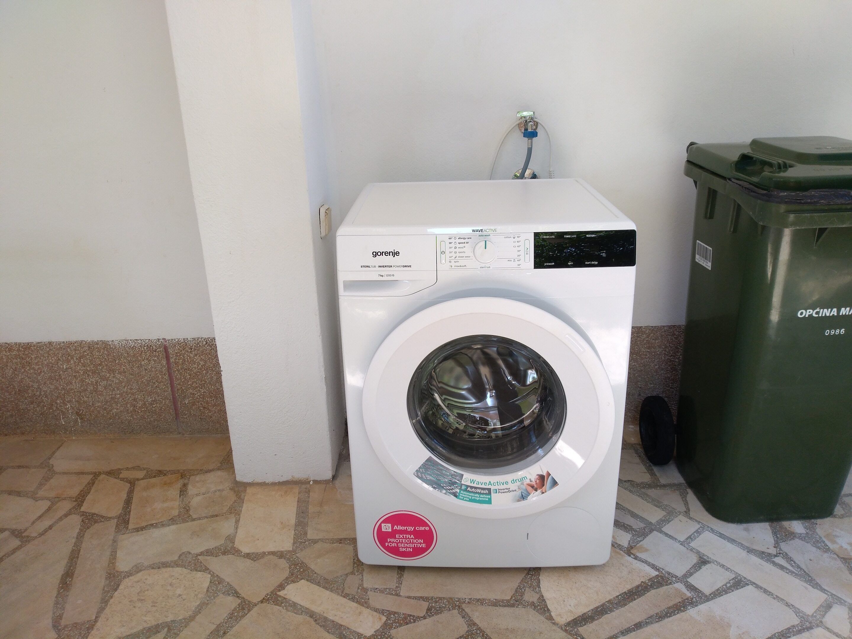 A white washing machine with a control panel, a water inlet, and a green trash bin beside it.
